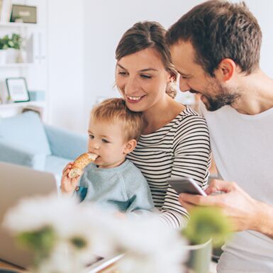 Eine glückliche Familie mit Mutter, Vater und Kind am Tisch vor einem Laptop und mit Smartphone