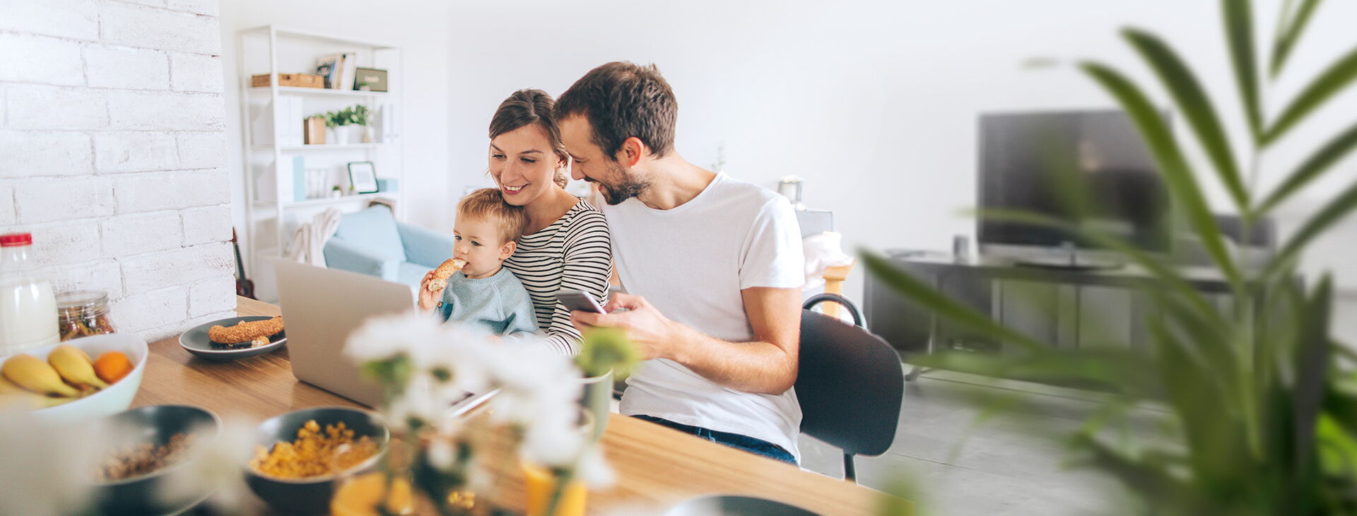 Eine glückliche Familie mit Mutter, Vater und Kind am Tisch vor einem Laptop und mit Smartphone