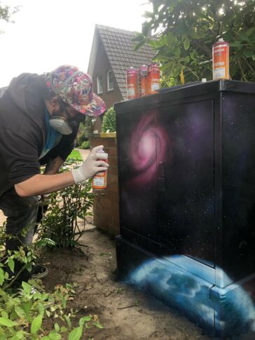 Ein Kabelverteiler-Schrank der von einer Person mit einem Galaxy Motiv besprüht wird