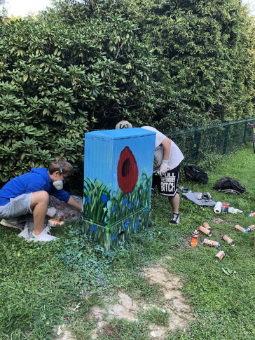 Zwei Personen wie sie Blumen und eine Wiese auf einen Kabelverteiler-Schrank malen