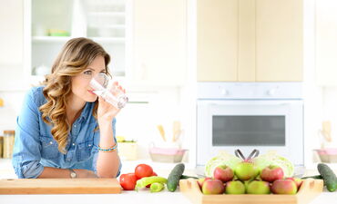 Frau steht in der Küche und trinkt ein Glas Wasser, daneben steht Obst