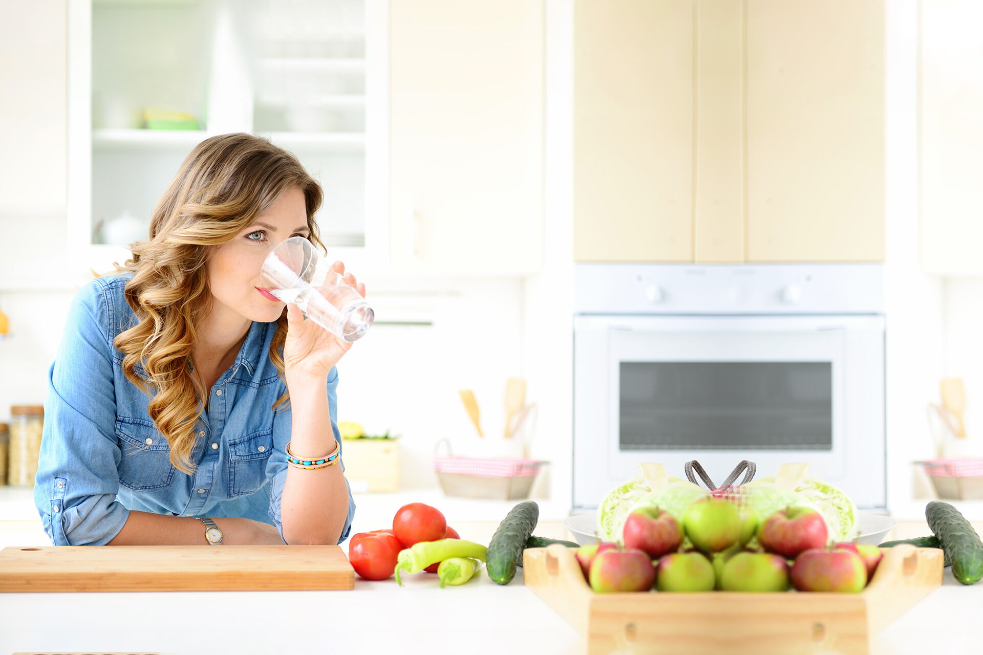 Frau steht in der Küche und trinkt ein Glas Wasser, daneben steht Obst