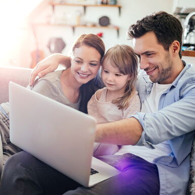 Mutter, Vater und Tochter liegen auf dem Sofa und schauen sich etwas auf dem Laptop an
